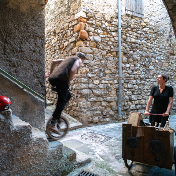 Image illustrant la version déambulatoire du spectacle. Dans une ruelle d'un village, lui descend des escaliers sur son monocycle. Elle pouse un cariole pleine d'accesoires