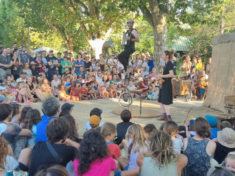 Image illustrant la version rue du spectacle. Les artistes sont entourés du public au festival de rue de valbonne. Lui joue un morceau d'accordeon sur un monocycle girafe, elle joue de la washboard.